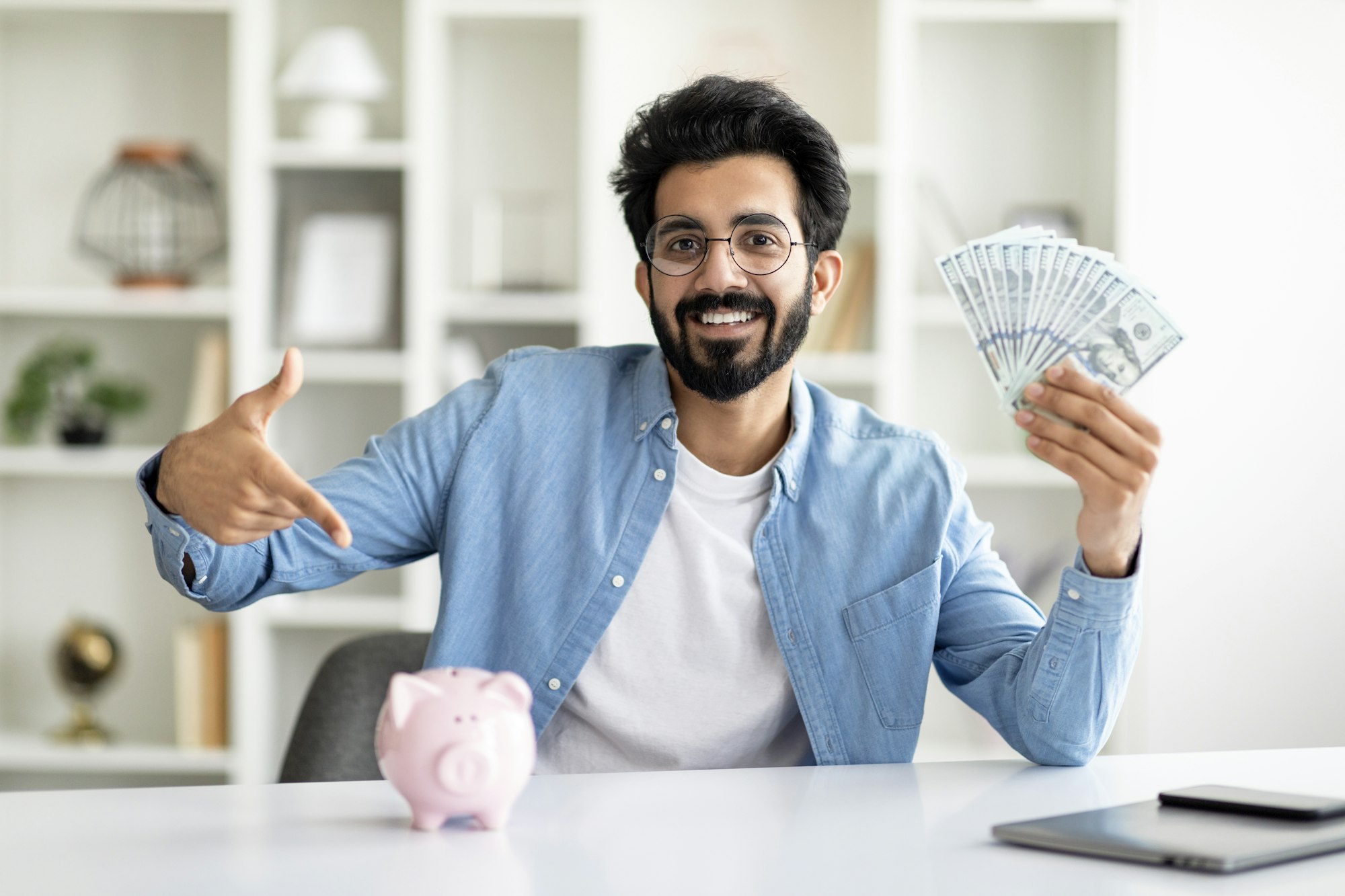 Handsome Indian Man Holding Dollar Cash And Pointing At Piggy Bank