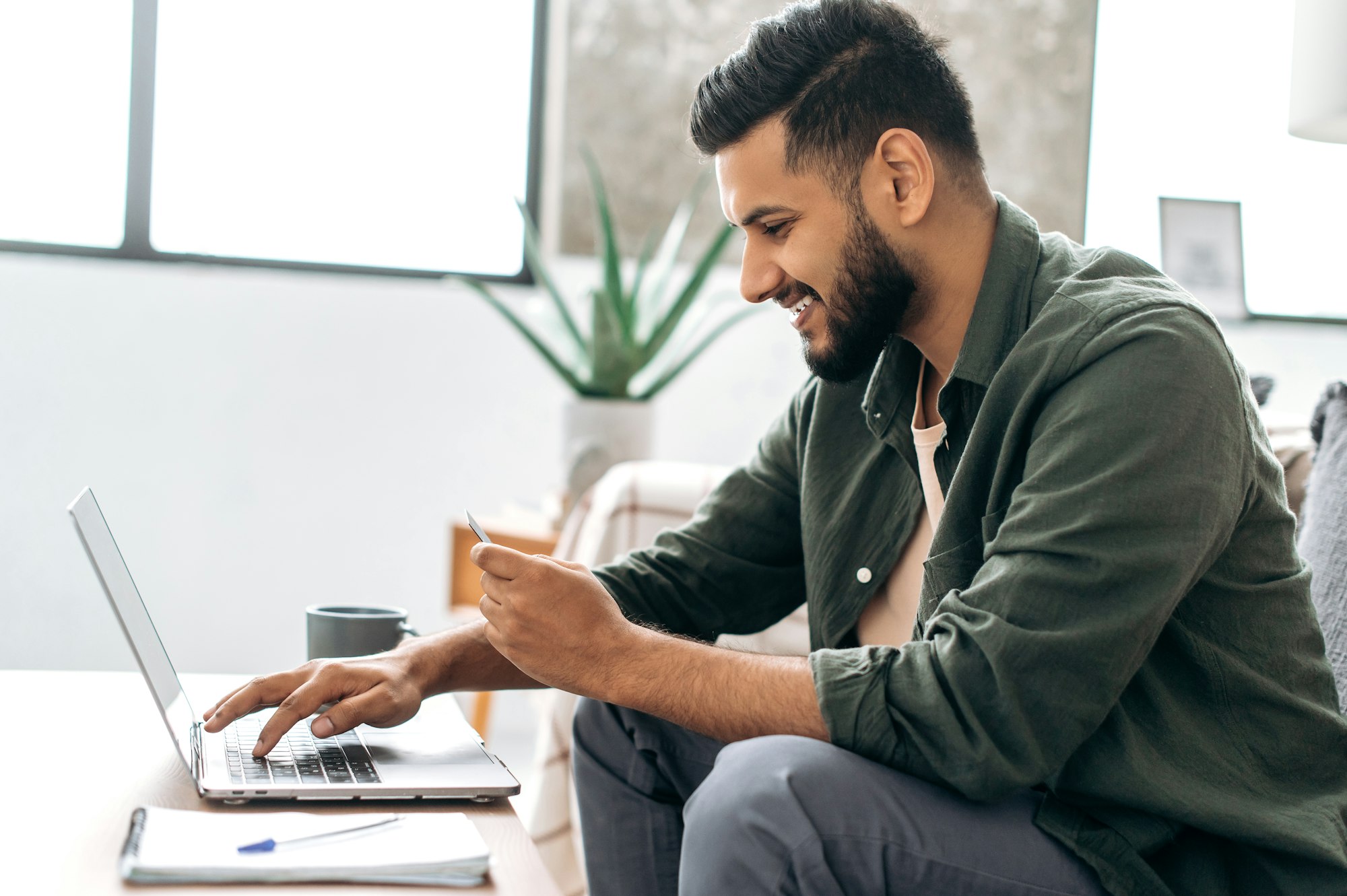 Side view of satisfied positive indian or arabian guy in stylish clothes sitting on a sofa in a